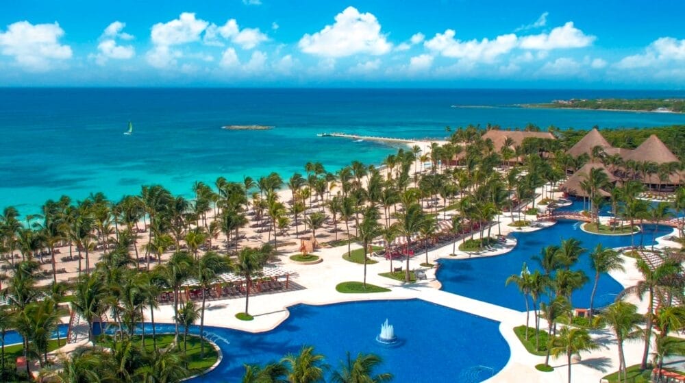 Vista aerea del Barcelo Maya Caribe, un resort Alpiselect con palme, grandi piscine, lettini ed edifici con tetti di paglia, che si affaccia sull'acqua turchese dell'oceano sotto un cielo blu brillante con nuvole sparse.
