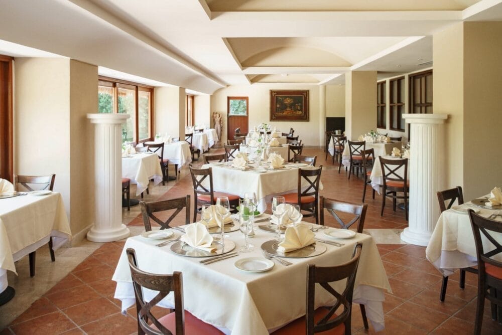 Elegante sala da pranzo del ristorante Allegro Playacar con tavoli rotondi ordinati, sedie in legno, tovaglie bianche, tovaglioli ripiegati, bicchieri da vino, ampie finestre, pavimento piastrellato e colonne classiche in un ambiente luminoso e spazioso.