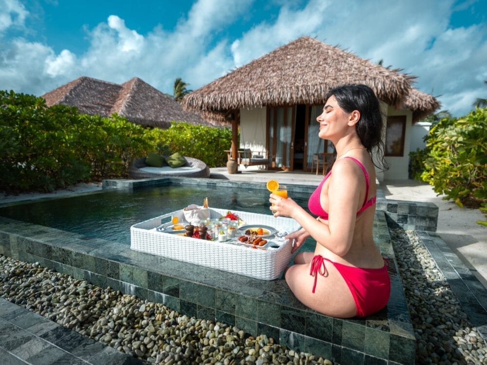 Una donna in bikini rosso siede a bordo di una piscina privata del Barceló Whale Lagoon, sorridendo e con un drink in mano, mentre davanti a lei galleggia un vassoio di cibo per la colazione. Sullo sfondo ville tropicali e vegetazione lussureggiante sotto un cielo azzurro.