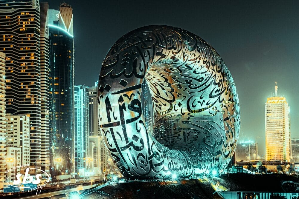 Un edificio futuristico di forma ovale con calligrafia araba sulla superficie metallica è illuminato di notte, circondato da moderni grattacieli: una tappa obbligata di qualsiasi tour di Dubai.