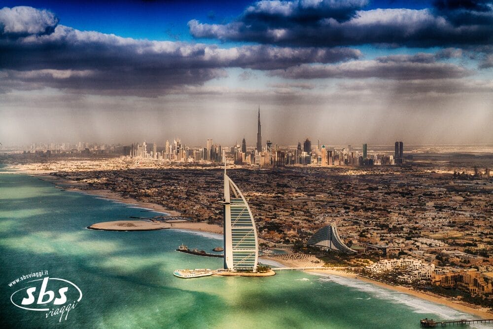 Veduta aerea della costa di Dubai con l'iconico hotel Burj Al Arab in primo piano e lo skyline della città, compreso il Burj Khalifa, sotto una drammatica coltre di nuvole sullo sfondo: una delle meraviglie del turismo nella regione emiratina.