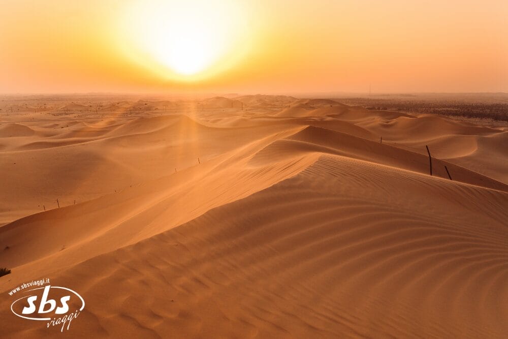 La luce dorata del sole illumina le vaste e ondulate dune di sabbia di un paesaggio desertico, evocando il fascino di un grand tour tra le sabbie degli Emirati. Il cielo limpido e il sole basso proiettano lunghe ombre e disegni ondulati. Nell'angolo in basso a sinistra compare un logo.