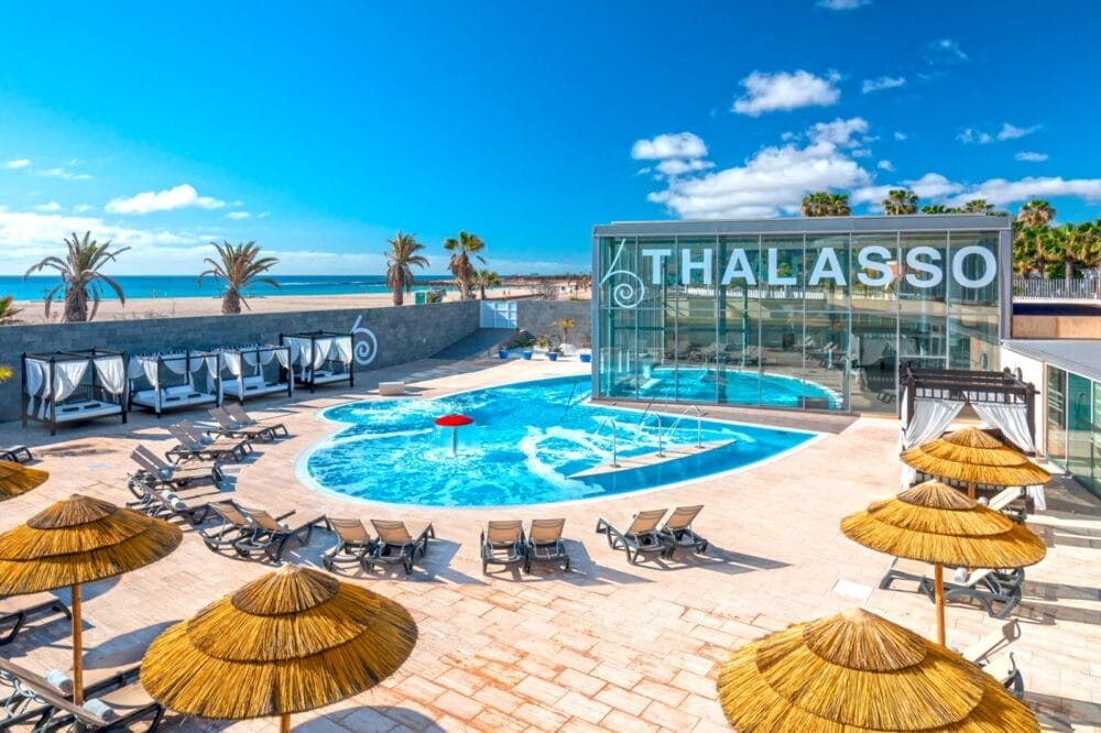 Area della piscina all'aperto del resort balneare Barcelo Fuerteventura con sedie a sdraio, ombrelloni di paglia e cabanas; un moderno edificio "Thalasso" in vetro e palme si affacciano sulla spiaggia e sull'oceano sotto un cielo azzurro.
