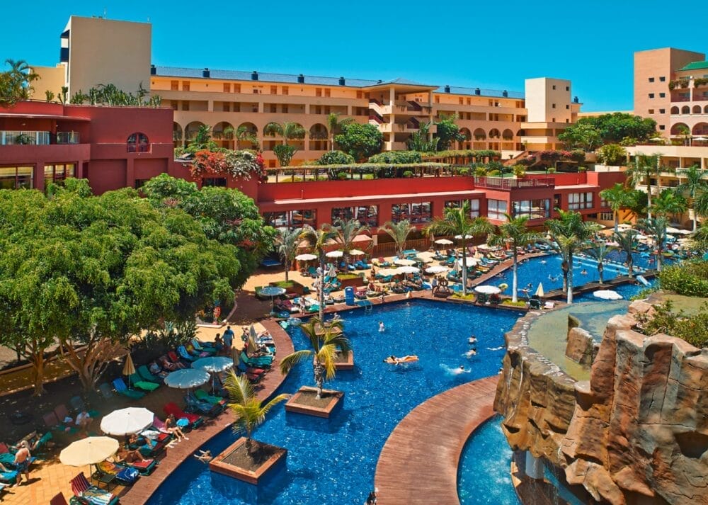 Il vivace resort Alpiclub dispone di una grande piscina all'aperto circondata da sedie a sdraio, palme e persone che si rilassano. L'edificio a più piani dell'hotel, incorniciato da alberi di jacaranda in fiore, si affaccia sulla piscina sotto un cielo blu brillante.