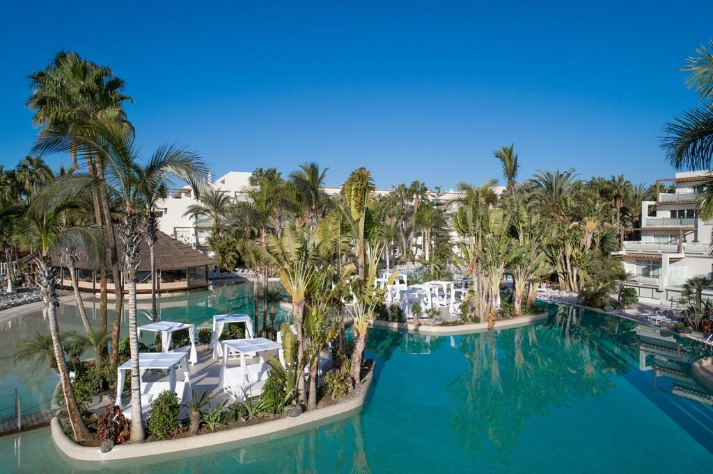 Una piscina del resort di Maspalomas circondata da palme e piante tropicali, con cabanas bianche su piccole isole e gli edifici di Maspalomas Princess sullo sfondo, sotto un cielo azzurro e limpido.