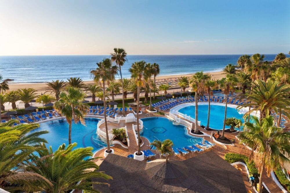 Un resort a Lanzarote con palme, due grandi piscine blu, molte sedie a sdraio e una spiaggia sabbiosa con le onde dell'oceano sullo sfondo di un cielo limpido.