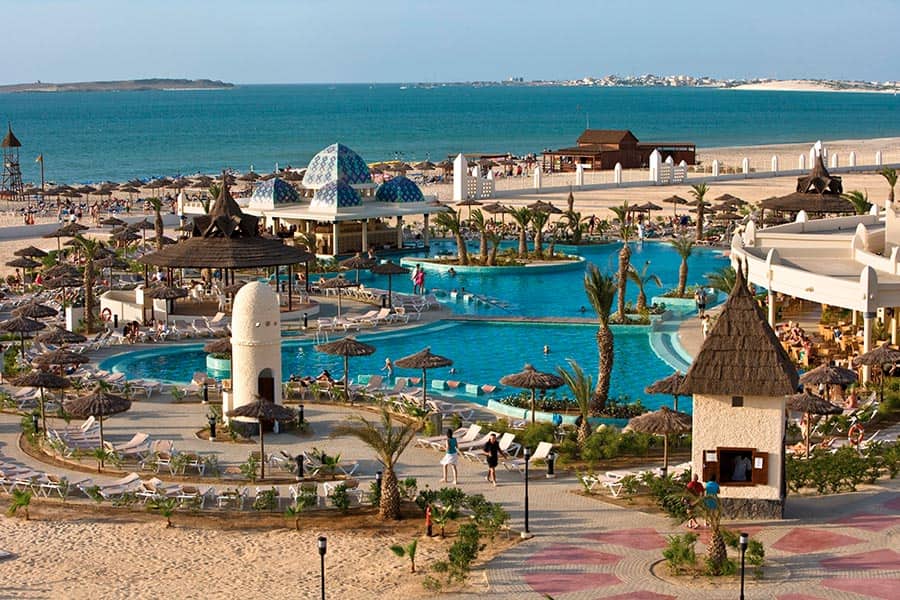 Un grande Searesort con piscine, sedie a sdraio, palme e cabine con tetto di paglia si affaccia su una spiaggia sabbiosa e sull'oceano blu sotto un cielo limpido. Persone che si rilassano in acqua e sulla spiaggia sullo sfondo del Riu Karamboa.