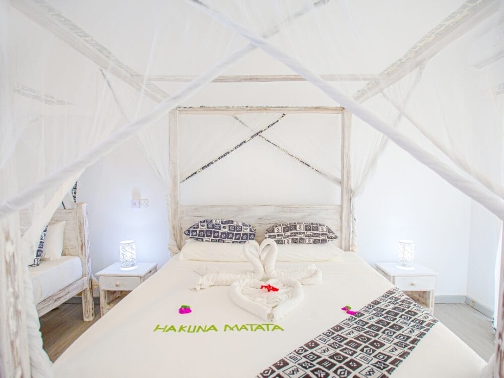 Al Lion Beach Resort vi attende un luminoso letto a baldacchino bianco con rete, cuscini a fantasia e un asciugamano piegato a forma di cigno sulla coperta. Le parole "Hakuna Matata" sono scritte in verde sul copriletto.