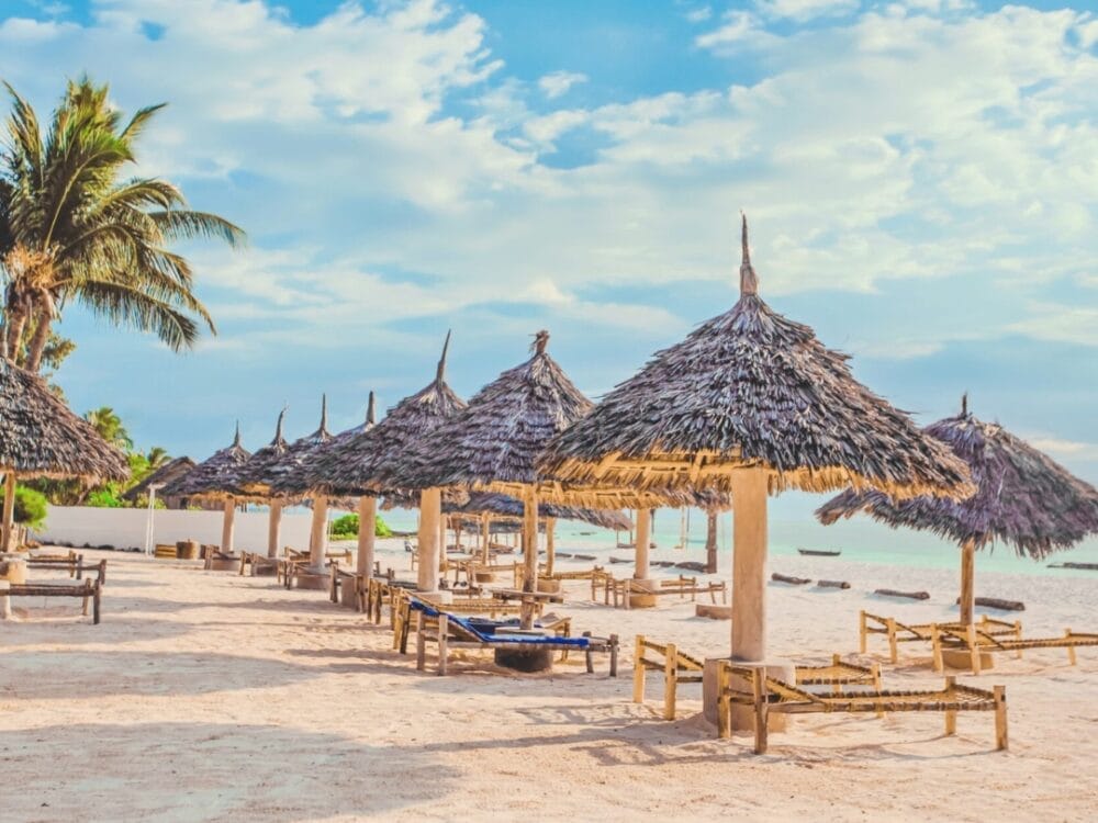Ombrelloni di paglia e sedie a sdraio in legno costeggiano la spiaggia sabbiosa del Waridi Beach Resort di Zanzibar, con palme sulla sinistra sotto un cielo parzialmente nuvoloso e un mare calmo e turchese.