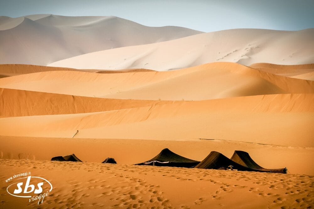 Paesaggio desertico con grandi dune di sabbia del Sahara sullo sfondo e diverse tende scure montate in primo piano, con deboli impronte sulla sabbia. Il logo "sbs image" è visibile nell'angolo in basso a sinistra, evocando una scena dell'avventura del Sahara Express.