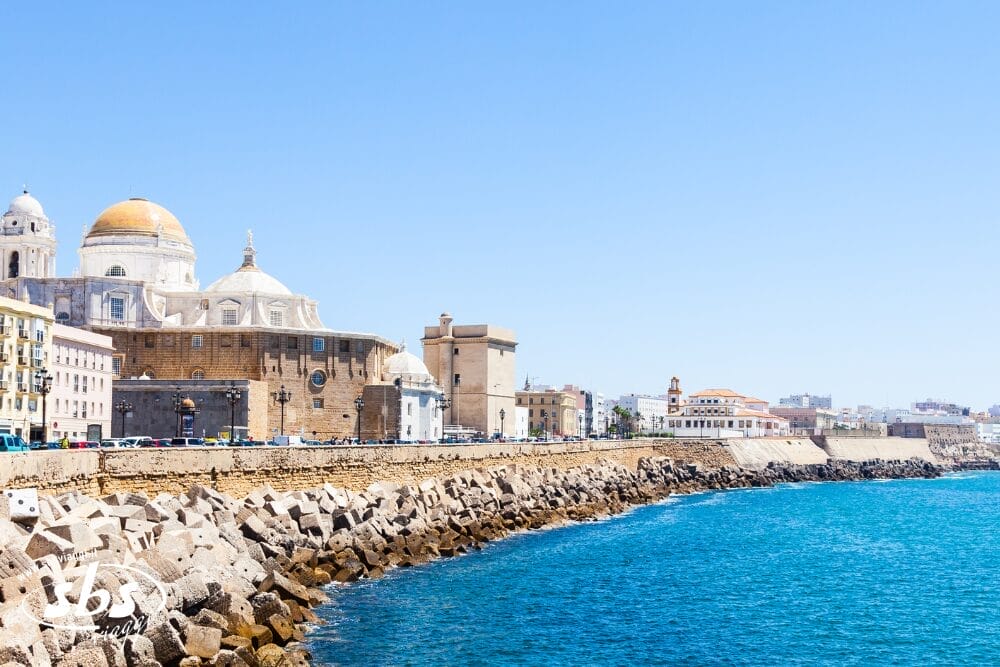 Vista della Cattedrale di Cádiz con il suo tetto a cupola, gli edifici storici e il lungomare in pietra lungo la costa blu brillante sotto un cielo limpido a Cádiz, in Spagna, una tappa essenziale di ogni Gran Tour Andalusia.
