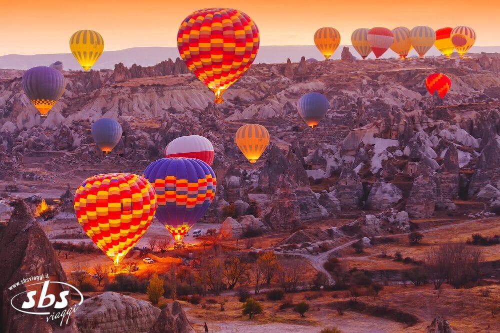 Colorate mongolfiere fluttuano sopra i paesaggi rocciosi e unici della Cappadocia all'alba durante il Grantour Turchia 2026. La luce calda illumina i palloni a grappolo che si alzano da terra, mentre altri si librano in alto nel cielo.