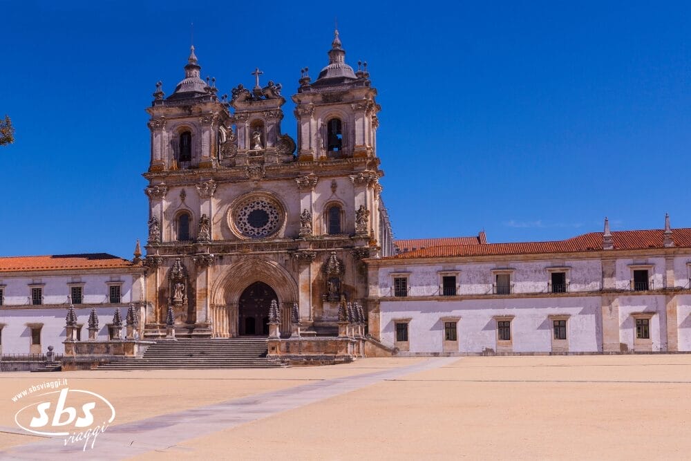 Uno storico monastero in pietra con due campanili gemelli e una facciata ornata si erge sotto un cielo azzurro e limpido a Portogallo. Il grande edificio ha porte ad arco, tetti di tegole rosse e un cortile aperto e sabbioso, perfetto per un Minitour del Portogallo.