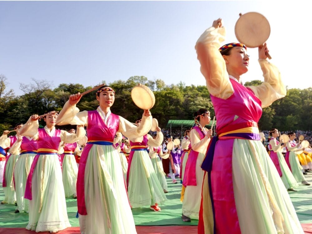 Un gruppo di donne in colorati abiti tradizionali coreani hanbok si esibisce in una danza del tamburo all'aperto, tenendo piccoli tamburi a mano e bacchette, celebrando il Sogno coreano sotto gli alberi e un cielo blu brillante della Corea del Sud.