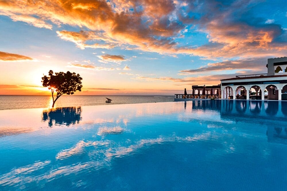 Una piscina a sfioro riflette il cielo vibrante del tramonto in questo hotel di lusso, con un albero solitario e vista sull'oceano. Un elegante edificio bianco con archi si trova sulla destra, mentre le nuvole sono illuminate dal sole che svanisce al Riu Palace Zanzibar.