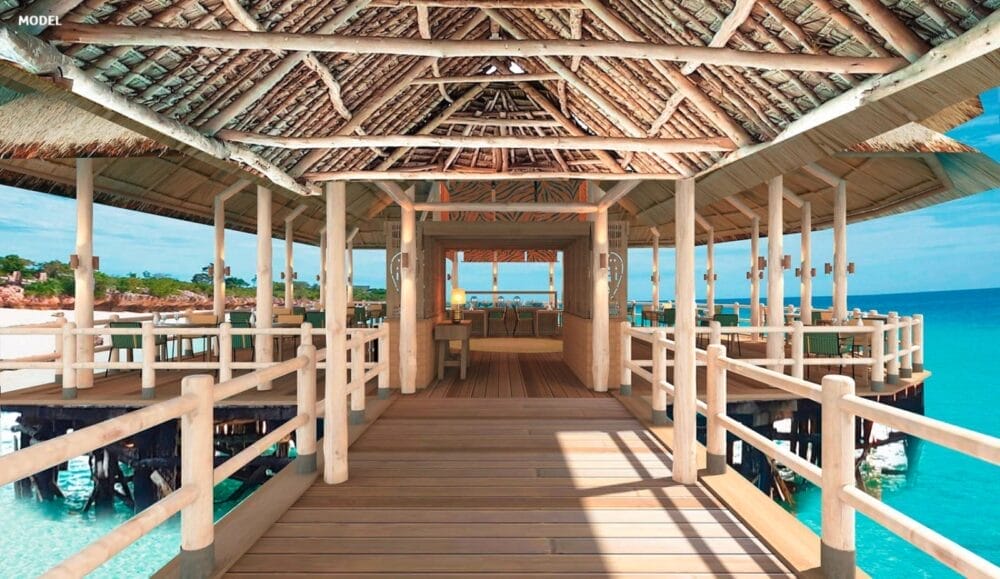 Un pontile di legno con un tetto di paglia conduce alla lounge all'aperto del Riu Jambo Hotel, che si affaccia sull'acqua turchese dell'oceano, con un cielo limpido e alberi verdi sullo sfondo.