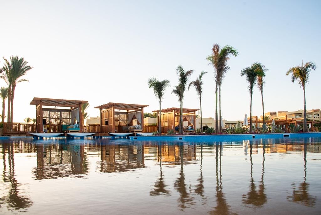 Una fila di cabine in legno con tende bianche si trova accanto a una piscina tranquilla, circondata da alte palme sotto il cielo azzurro di un resort.
