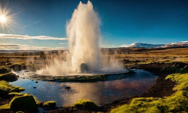 Un geyser erutta, sparando acqua e vapore in aria, circondato da erba verde e da una piscina riflettente sotto un sole splendente. Le montagne innevate e lo spettacolo impressionante di una Bozza automatica completano la scena mozzafiato.