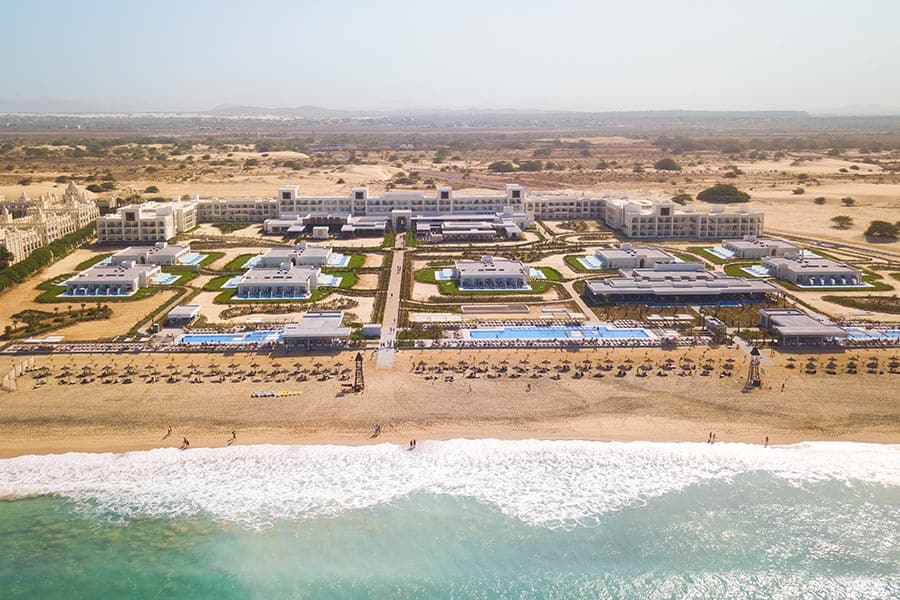Vista aerea del Riu Palace Boavista, un grande resort sulla spiaggia con diverse piscine, prati verdi ed edifici bianchi, situato su un terreno sabbioso vicino all'oceano, con le onde che colpiscono dolcemente la riva.