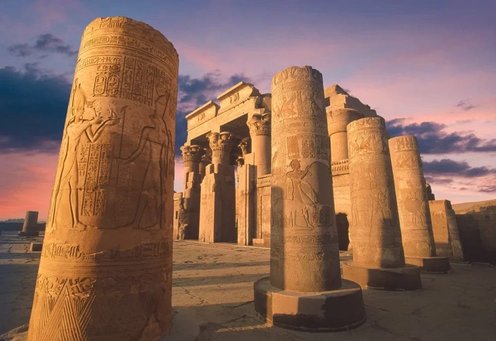 Antiche colonne di pietra con geroglifici e incisioni si ergono sotto il cielo colorato del tramonto al Tempio di Kom Ombo, un'attrazione spesso visitata durante la Crociera Ramses dopo aver esplorato le meraviglie di Luxor, con le rovine e le drammatiche nuvole sullo sfondo.