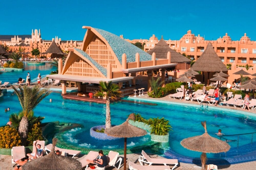 Un resort tropicale con una grande piscina all'aperto, un bar, palme, lettini e persone che si rilassano sotto ombrelloni di paglia. Edifici arancioni dall'architettura unica fanno da sfondo a un cielo azzurro e limpido.