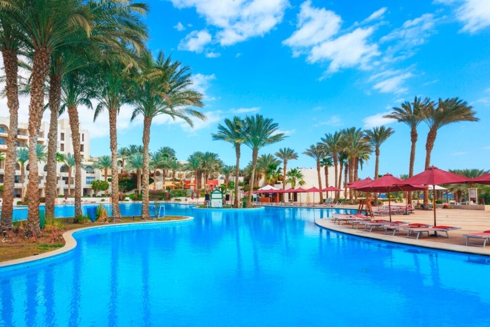 Una grande piscina all'aperto del Seaclub Grand Rotana Resort, circondata da palme, sedie a sdraio con ombrelloni rossi ed edifici del resort, sotto un cielo blu brillante cosparso di nuvole.