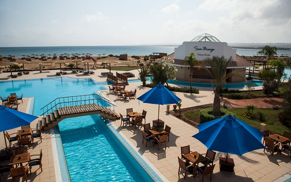 Area della piscina all'aperto del Seaclub Lahami Bay con tavoli in legno, ombrelloni blu, passerella sulla piscina, palme, edificio del ristorante e vista sul mare, sulla spiaggia sabbiosa e sui lettini sullo sfondo.