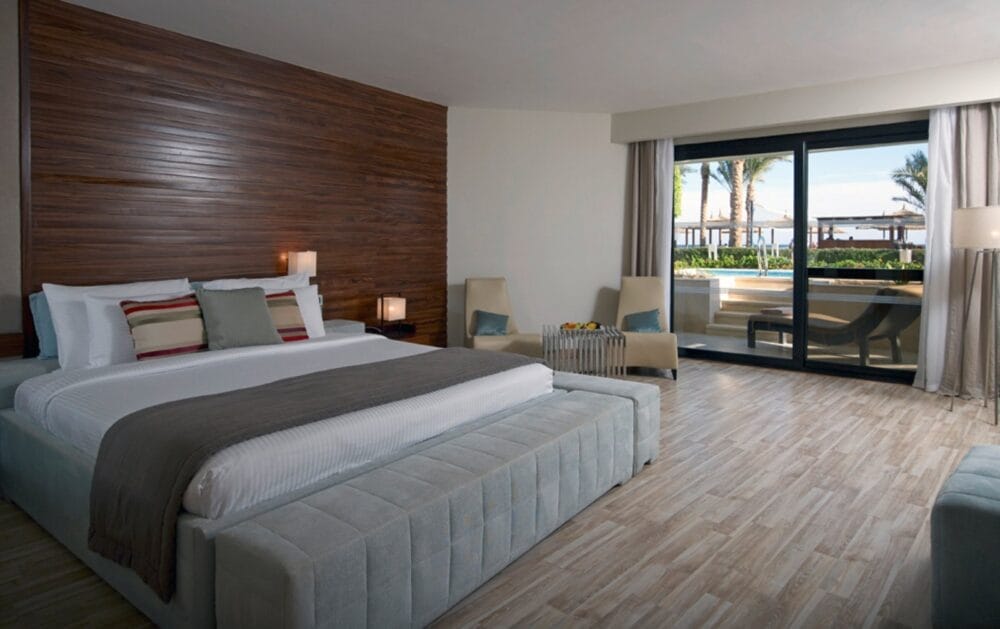 Una moderna camera d'albergo del Sensatori con un grande letto, una parete con accenti in legno, un'accogliente area salotto e porte scorrevoli in vetro che si aprono su un patio con sedie a sdraio e vista mozzafiato sulle palme e sul Mar dei Coralli Imperiale.