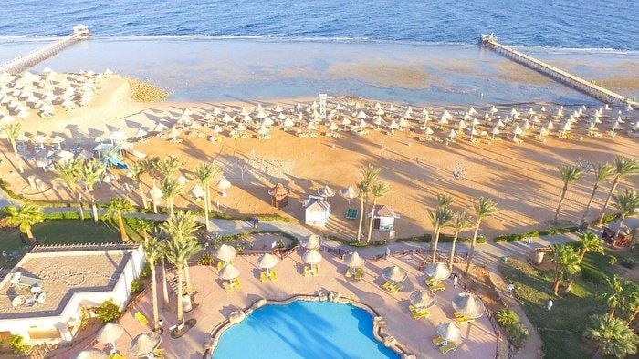 Vista aerea del Parrotel Beach Resort di Sharm El Sheikh, con file di ombrelloni sulla sabbia, palme, una piscina e due moli che si estendono nel mare blu.