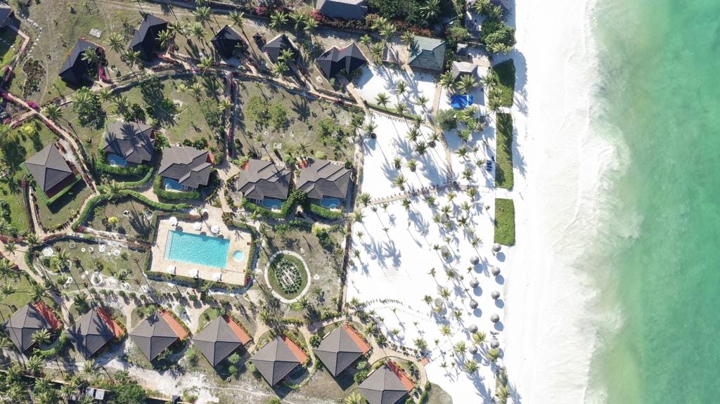 Vista aerea di un resort sulla spiaggia con ville, piscina, palme, spiaggia di sabbia bianca, ombrelloni di paglia e acqua turchese lungo la costa: la vostra perfetta fuga automatica di Bozza.