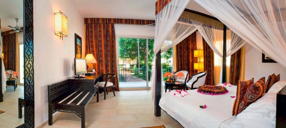 La camera d'albergo, luminosa ed elegantemente arredata in stile Tui Blue Bahari, dispone di un letto a baldacchino da sogno ornato di petali di fiori, cuscini a motivi colorati, una scrivania, un grande specchio e porte scorrevoli in vetro che conducono al patio con vista sul giardino.