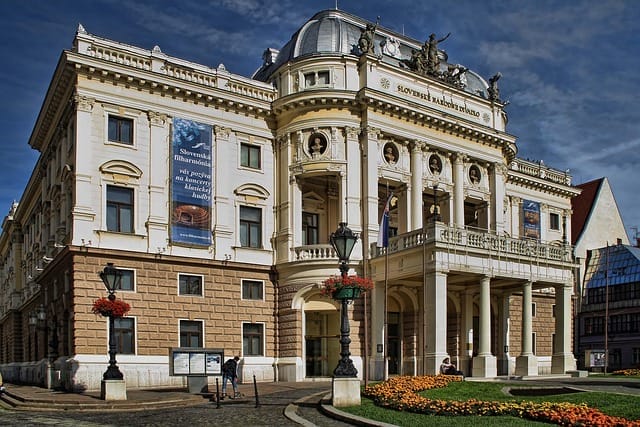 Edificio storico con colonne ornate e statue sul tetto, il Teatro Nazionale Slovacco di Bratislava evoca la grandezza dei Capitali Imperiali di Europa Centrale, con i suoi stendardi, le finestre ad arco, il tetto a cupola, i lampioni e le vivaci aiuole.