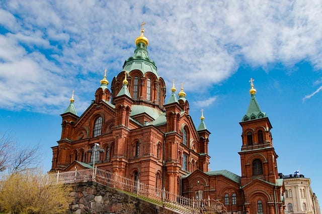 Una grande cattedrale in mattoni rossi con tetti verdi e cupole dorate si staglia contro un cielo blu brillante a Helsinki, circondata da una recinzione e da alberi in primo piano.