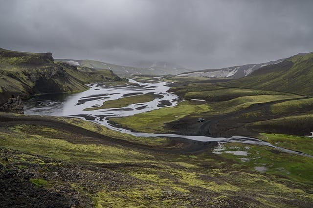 Un fiume sinuoso attraversa un paesaggio vulcanico verde e nero sotto un cielo nuvoloso e coperto, con colline ondulate e macchie di neve sullo sfondo: una scena che ricorda un capolavoro dell'automatismo di Bozza.
