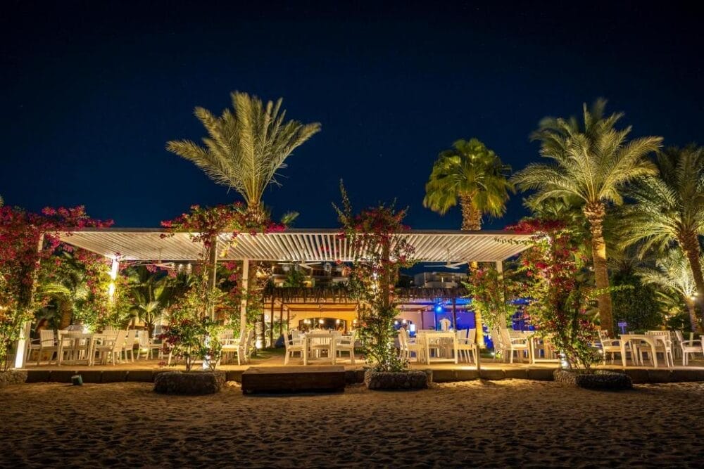 Un ristorante all'aperto di notte, circondato da palme e piante fiorite al Fort Arabesque Resort, con tavoli e sedie bianche sotto un pergolato, illuminato da un cielo scuro.