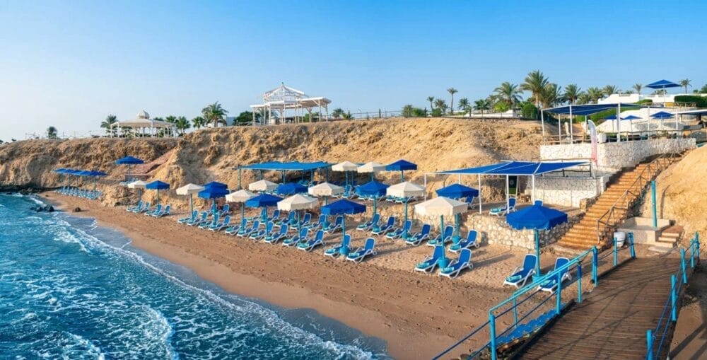File di lettini e ombrelloni blu costeggiano l'arenile del Reef Beach Resort accanto all'acqua cristallina, con scogliere rocciose e palme sullo sfondo sotto un cielo limpido. I gradini scendono dalla terrazza del Bravo Smart Club.