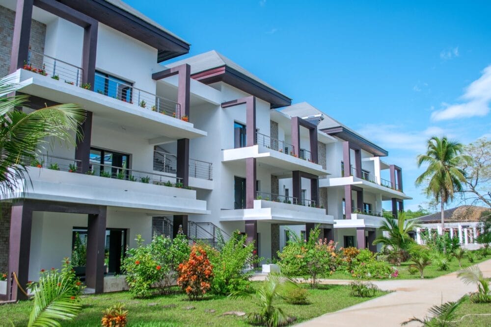 Il moderno condominio bianco con ampi balconi, circondato da piante tropicali e palme, offre un'atmosfera da resort che ricorda il Seaclub Royal Andilana Resort. Un sentiero lastricato si snoda davanti al lussureggiante giardino paesaggistico sotto un cielo blu brillante.