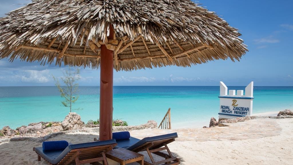Due sedie a sdraio in legno con cuscini si trovano sotto un ombrellone di paglia sulla spiaggia sabbiosa del Royal Zanzibar Beach Resort, di fronte all'acqua turchese dell'oceano. Un cartello indica il resort vicino a gradini di pietra che conducono alla riva per il massimo relax.