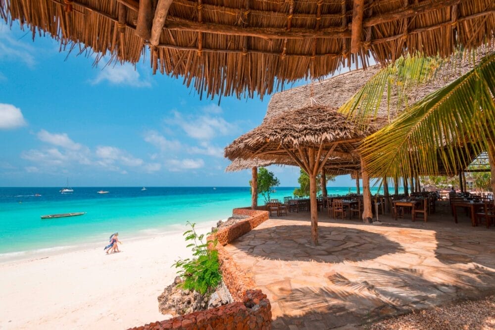 Il ristorante tropicale sulla spiaggia del Seaclub Sandies Baobab si affaccia sull'acqua turchese e sulla sabbia bianca di questo invitante resort sulla spiaggia di Zanzibar. Tavoli di legno ombreggiati da fronde di palma attendono sotto un cielo azzurro e limpido, mentre poche persone passeggiano lungo la riva.