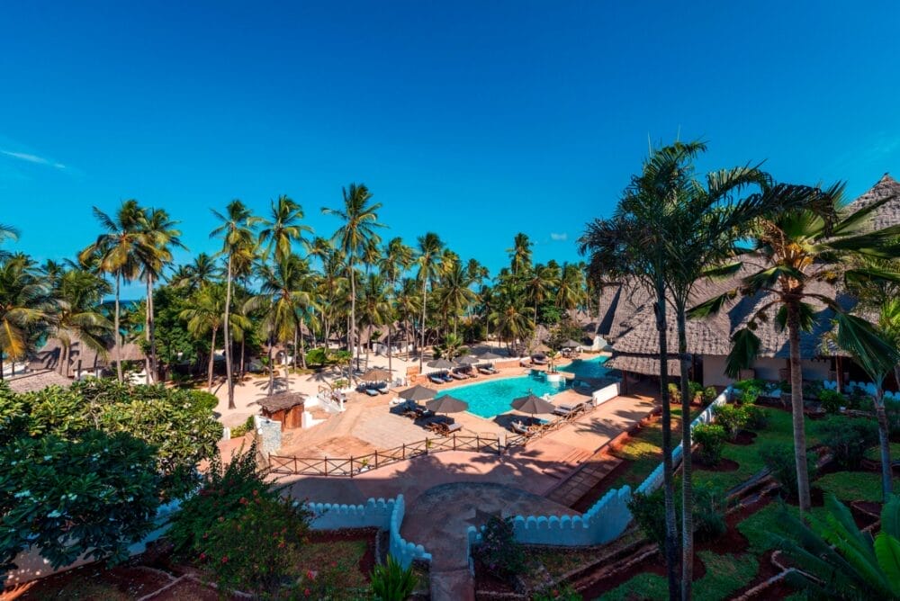 Un resort tropicale con edifici dai tetti di paglia, una piscina turchese e lettini circondati da alte palme sotto un cielo azzurro e limpido, il Seaclub Diamond Mapenzi Beach offre un ambiente lussureggiante e invitante per una vacanza all'insegna del relax.