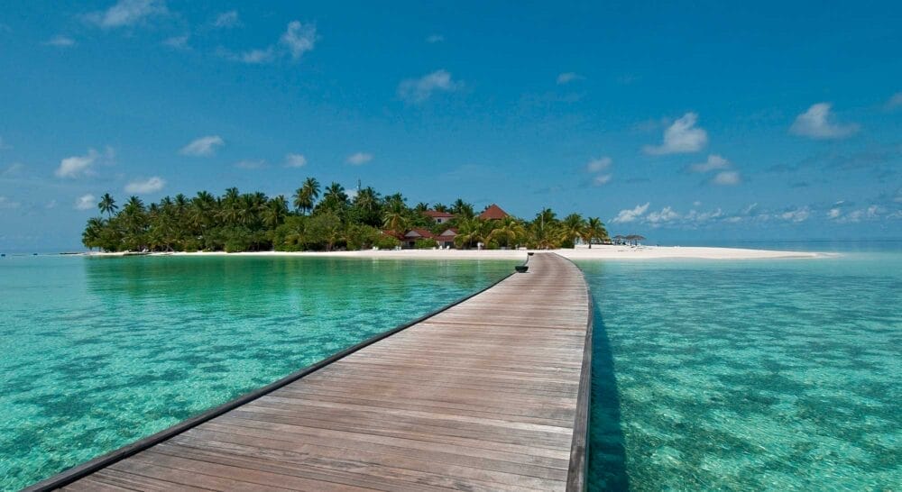 Una lunga passerella di legno si estende su un'acqua turchese e limpida verso un'isola tropicale lussureggiante con palme e spiagge di sabbia bianca, che conduce a una Water Villa sotto un cielo blu brillante con nuvole sparse, evocando il vero stile Seaclub.
