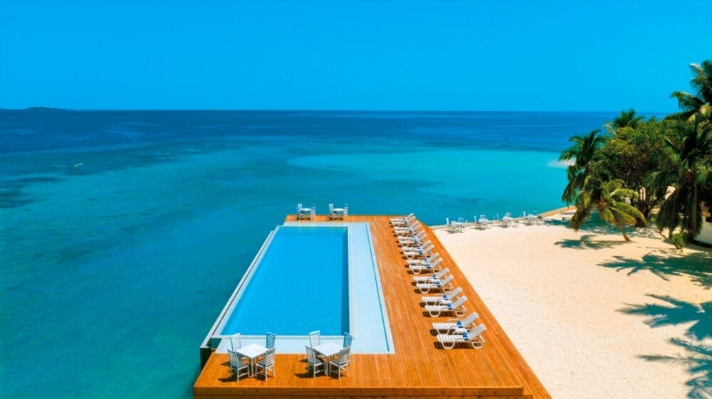 Una piscina a sfioro con sedie a sdraio su un ponte di legno si affaccia su una spiaggia sabbiosa e sull'oceano cristallino del Sandies Bathala Resort, delimitato da palme sotto un cielo blu brillante.