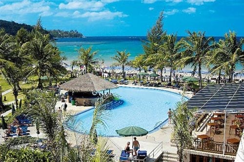 La piscina del Sunprime Kamala Beach Resort, circondata da palme, si affaccia sul litorale sabbioso e sull'oceano turchese di Kamala Beach, con persone che si rilassano in acqua e sotto gli ombrelloni sotto un cielo blu brillante.