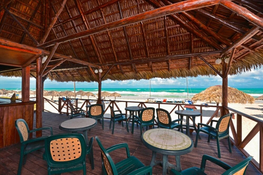 Un patio ombreggiato sulla spiaggia dell'Iberostar Tainos Seaclub presenta tavoli e sedie vuote e si affaccia su una spiaggia sabbiosa punteggiata di ombrelloni di paglia e sull'acqua turchese dell'oceano sotto un cielo parzialmente nuvoloso.