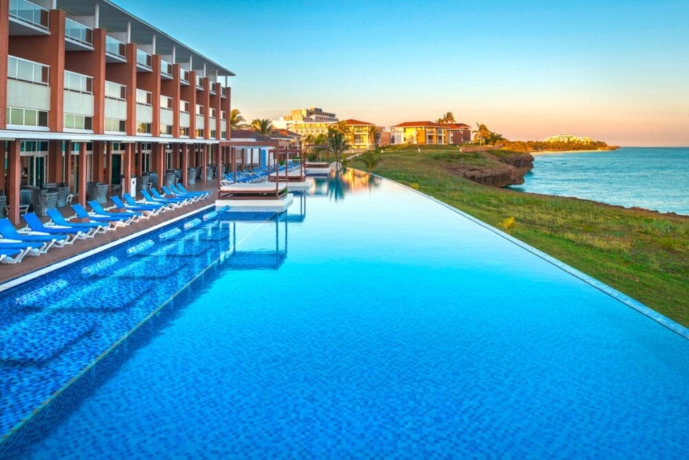 Una grande piscina a sfioro accanto al moderno hotel Playa Vista Azul con sedie a sdraio blu, con vista sull'oceano al tramonto. Sullo sfondo sono visibili altri edifici del Searesort, situati su un terreno erboso in riva al mare.