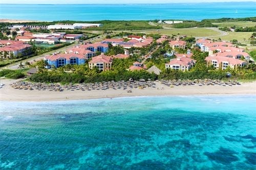 La veduta aerea del Searesort Iberostar Playa Alameda mostra edifici dai tetti rossi, palme e file di ombrelloni su una spiaggia sabbiosa accanto a un'acqua limpida e turchese, con un paesaggio verde sullo sfondo.