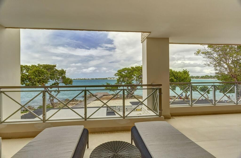 Un balcone coperto con due sedie a sdraio del Royalton Negril si affaccia sull'oceano, sulla spiaggia sabbiosa e sugli alberi verdi sotto un cielo parzialmente nuvoloso. La vista è tranquilla e invitante, e promette un rilassante ritiro in riva al mare in questo resort di lusso.