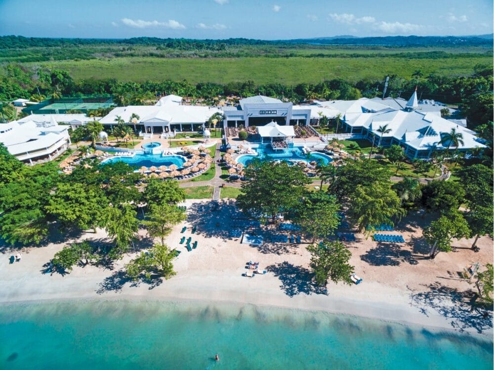 Vista aerea del Seaclub Riu Negril, uno splendido hotel della Giamaica con edifici bianchi, piscine, palme, lettini e ombrelloni accanto a una spiaggia sabbiosa e un'acqua cristallina, il tutto circondato da una vegetazione lussureggiante.