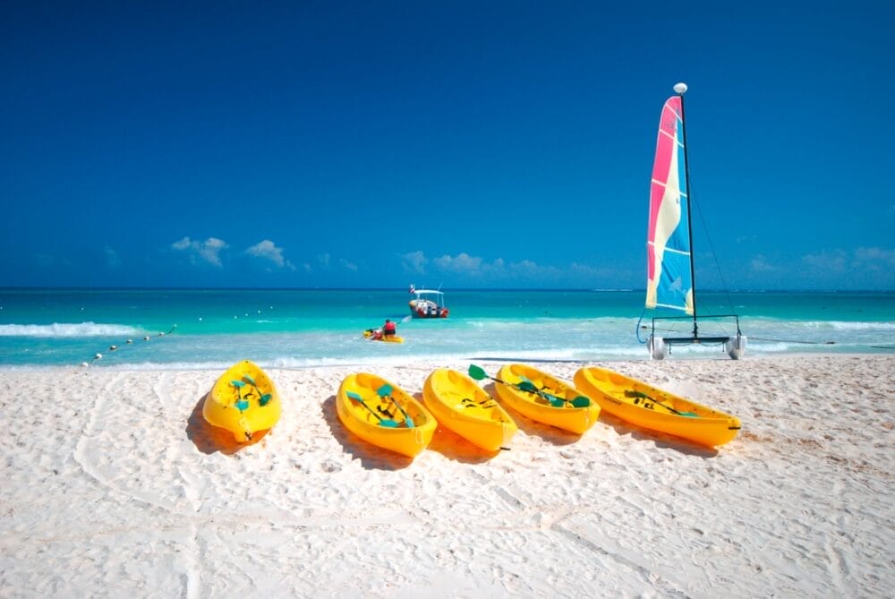Kayak gialli e colorati e una barca a vela costeggiano la spiaggia di sabbia bianca di Catalonia Playa Maroma, con l'acqua turchese dell'oceano e un cielo azzurro e limpido, con persone che fanno kayak e una barca in lontananza.