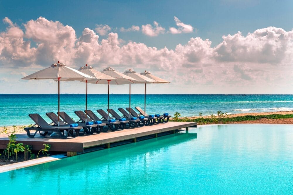 Una fila di sedie a sdraio con ombrelloni bianchi si estende su un ponte di legno accanto alla tranquilla piscina dell'Ocean Riviera Paradise Searesort, che si affaccia su una spiaggia sabbiosa e sull'oceano turchese sotto un cielo parzialmente nuvoloso.